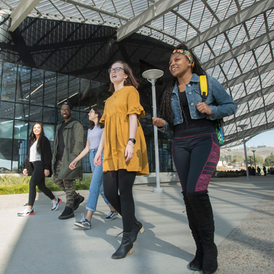 Five university students walking underneath glass-covered section of campus-area building