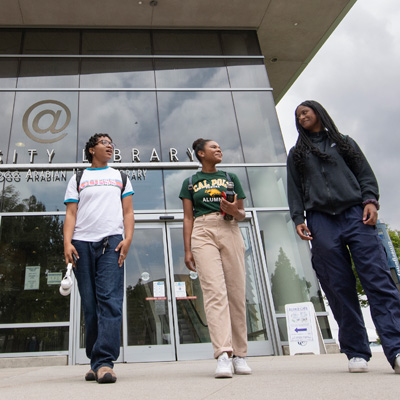Three university students walking near glass-covered entrance of campus-area library building