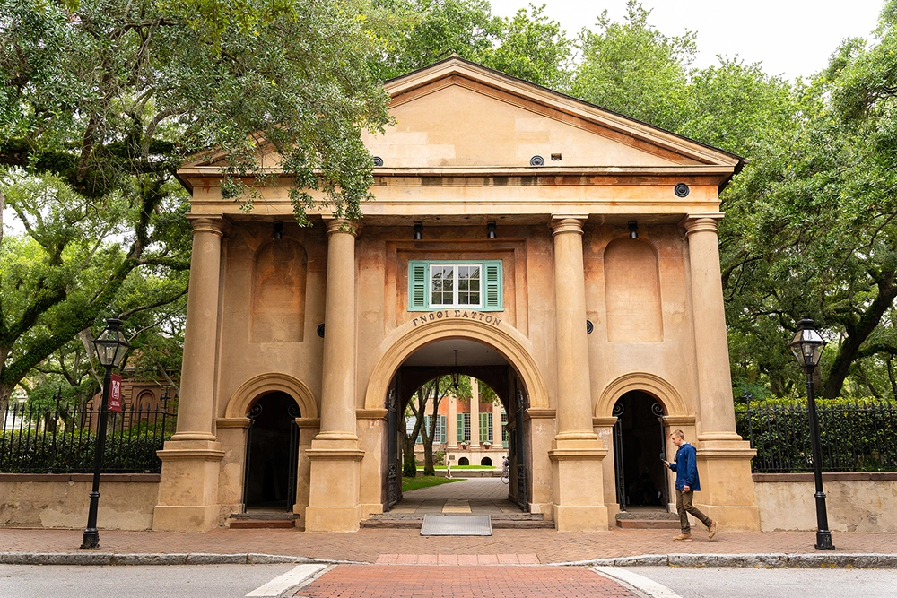 Student walking on brick sidewalk in front of archway facade of old university building nestled under trees