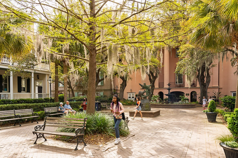 People walking and sitting in tree-shaded plaza among benches and fountains near campus buildings