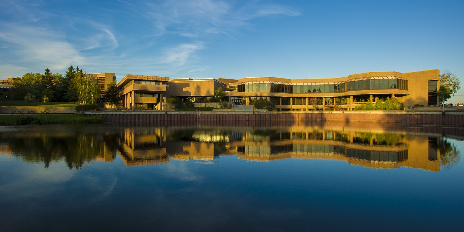 Panorama of Lakehead University campus in Ontario, Canada, with University Centre building reflected in calm waters of Lake Tamblyn