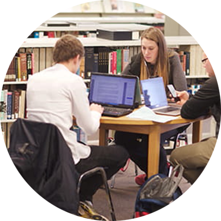 Circular image depicting students seated at table working on laptops among bookshelves in university library