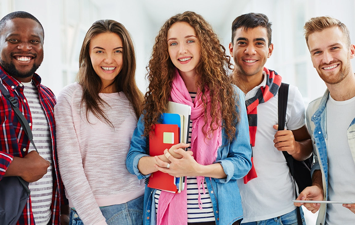 Five casually dressed university students smile into the camera while holding backpacks, textbooks or tablet devices