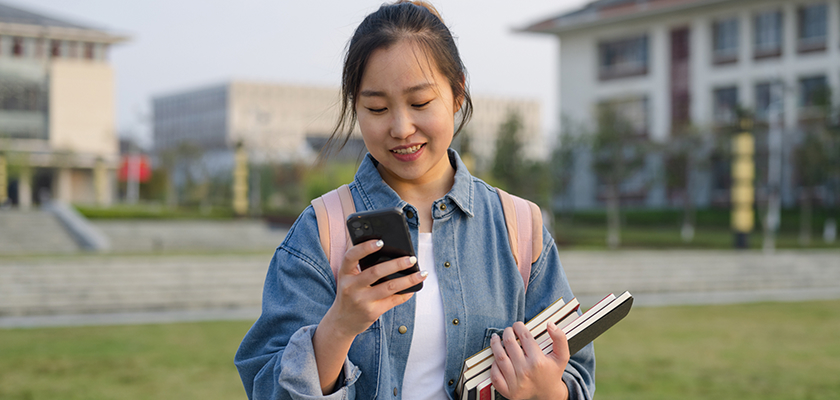 A student holds books in one hand and looks at her mobile device in the other hand.