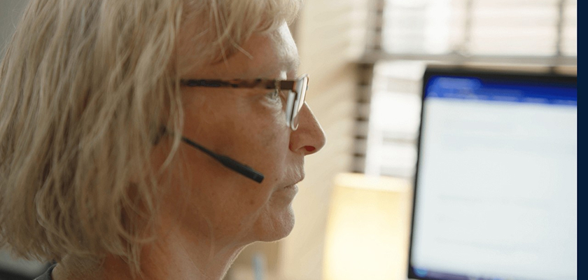 A closeup of an administrator wearing glasses and a headset microphone with a blurred computer screen in the back.
